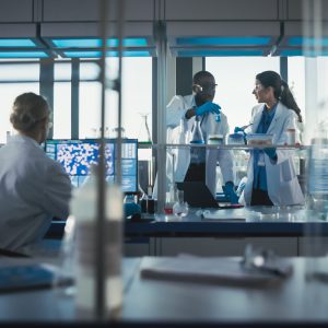 Scientists wearing lab coats and safety goggles working collaboratively in a modern laboratory with advanced equipment and computers in the background.